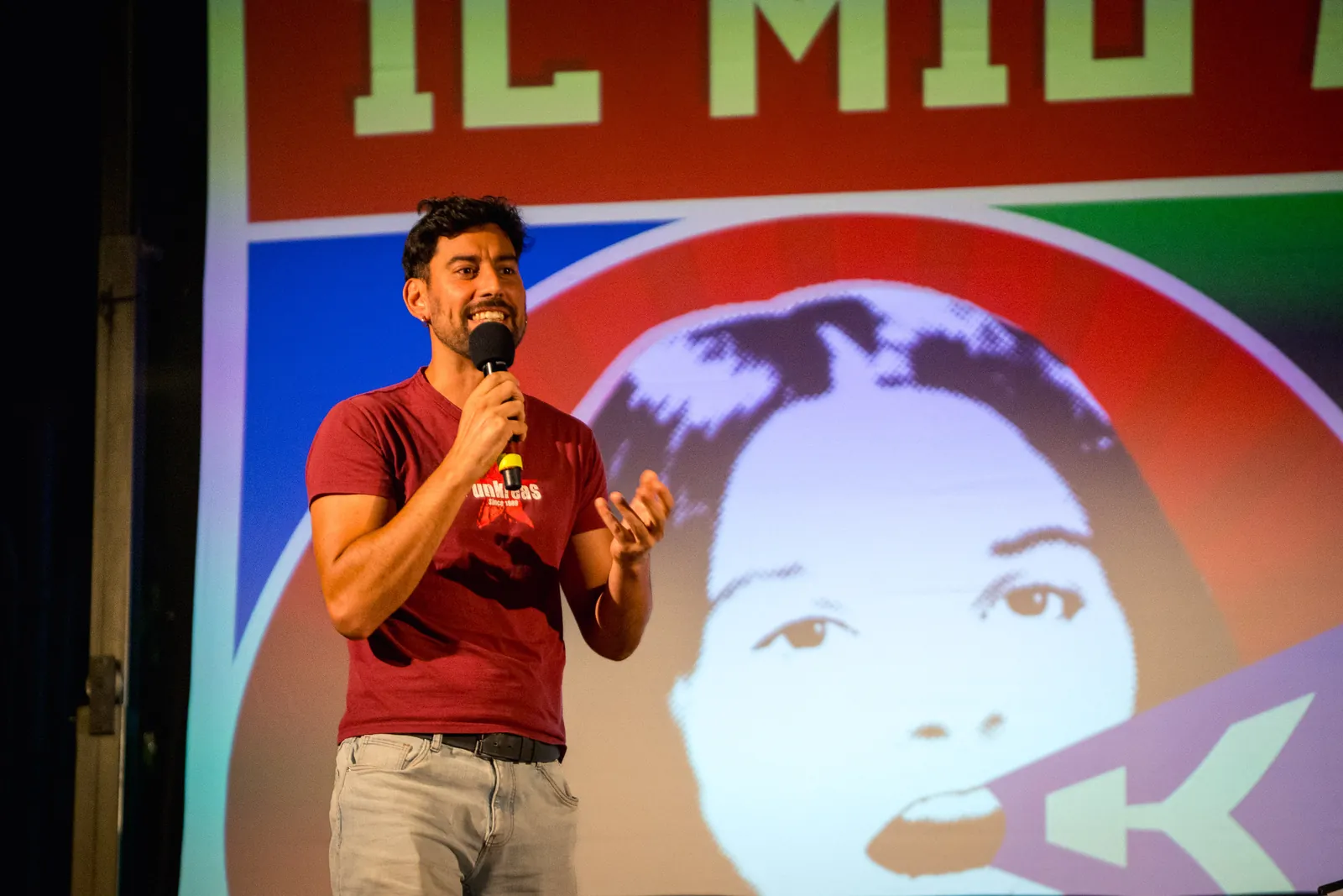 Un uomo in maglietta rossa parla al microfono sul palco, con un fotogramma pop-art del cortometraggio proiettato sullo schermo alle sue spalle