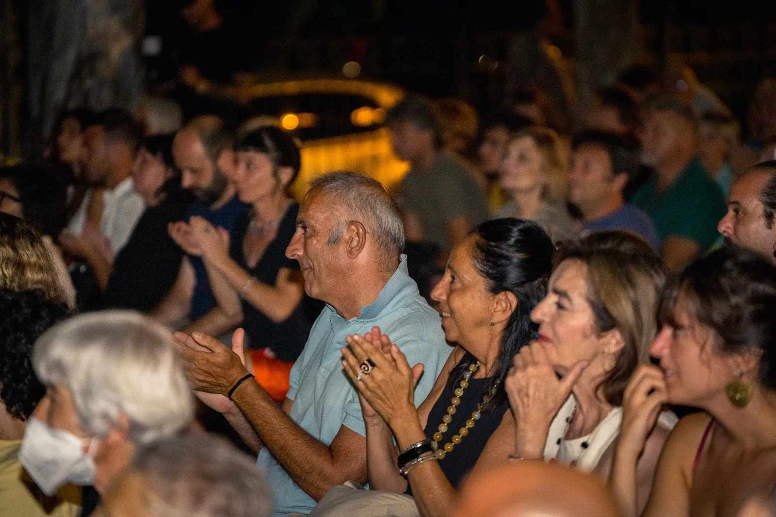 Il pubblico applaude in prima fila, con una coppia di spettatori sorridenti sotto le luci serali