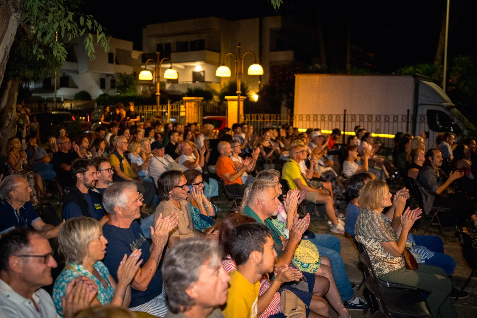 Il pubblico numeroso ripreso da dietro, seduto tra gli alberi con i lampioni e le palazzine sullo sfondo