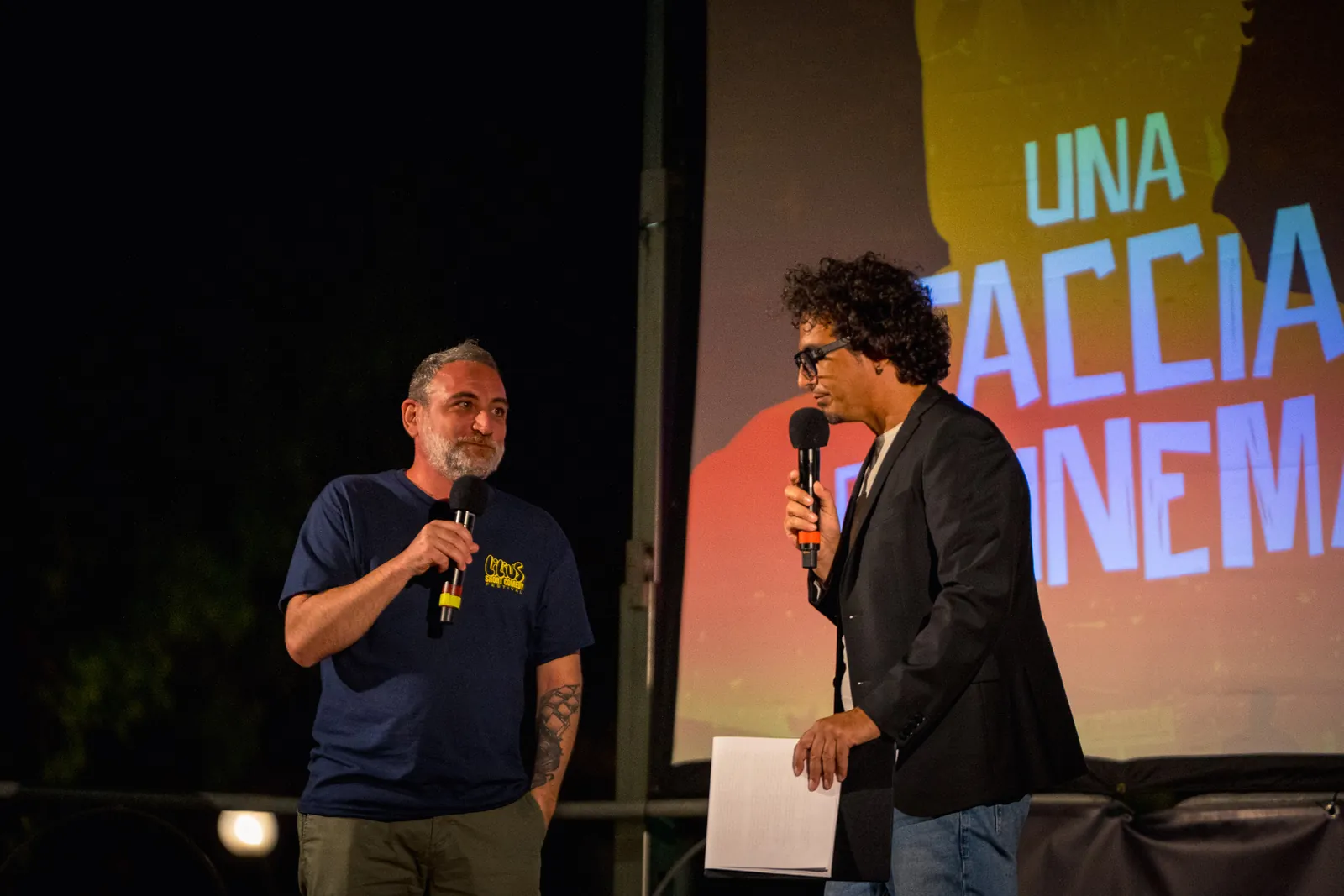 Due uomini sul palco con i microfoni, con il titolo del cortometraggio Una faccia da cinema proiettato sullo schermo alle spalle