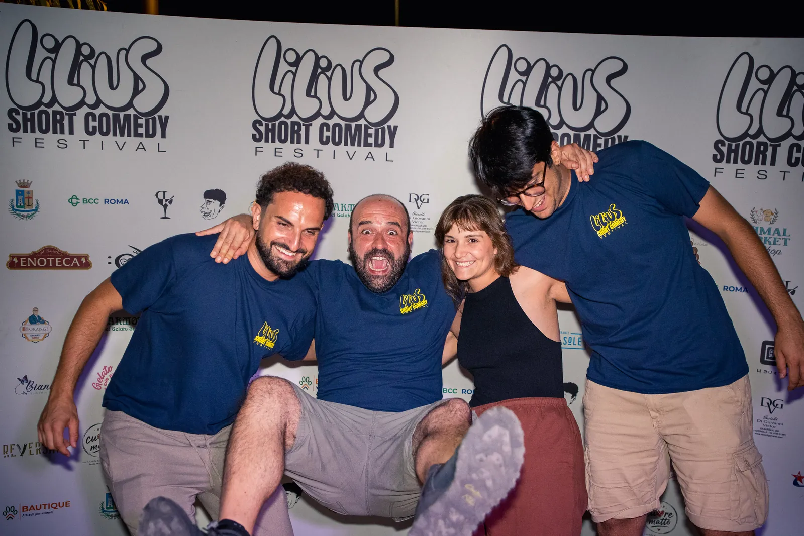 Un gruppo di quattro amici abbracciati e sorridenti al photo-wall del festival, in posa scherzosa con uno sollevato da terra