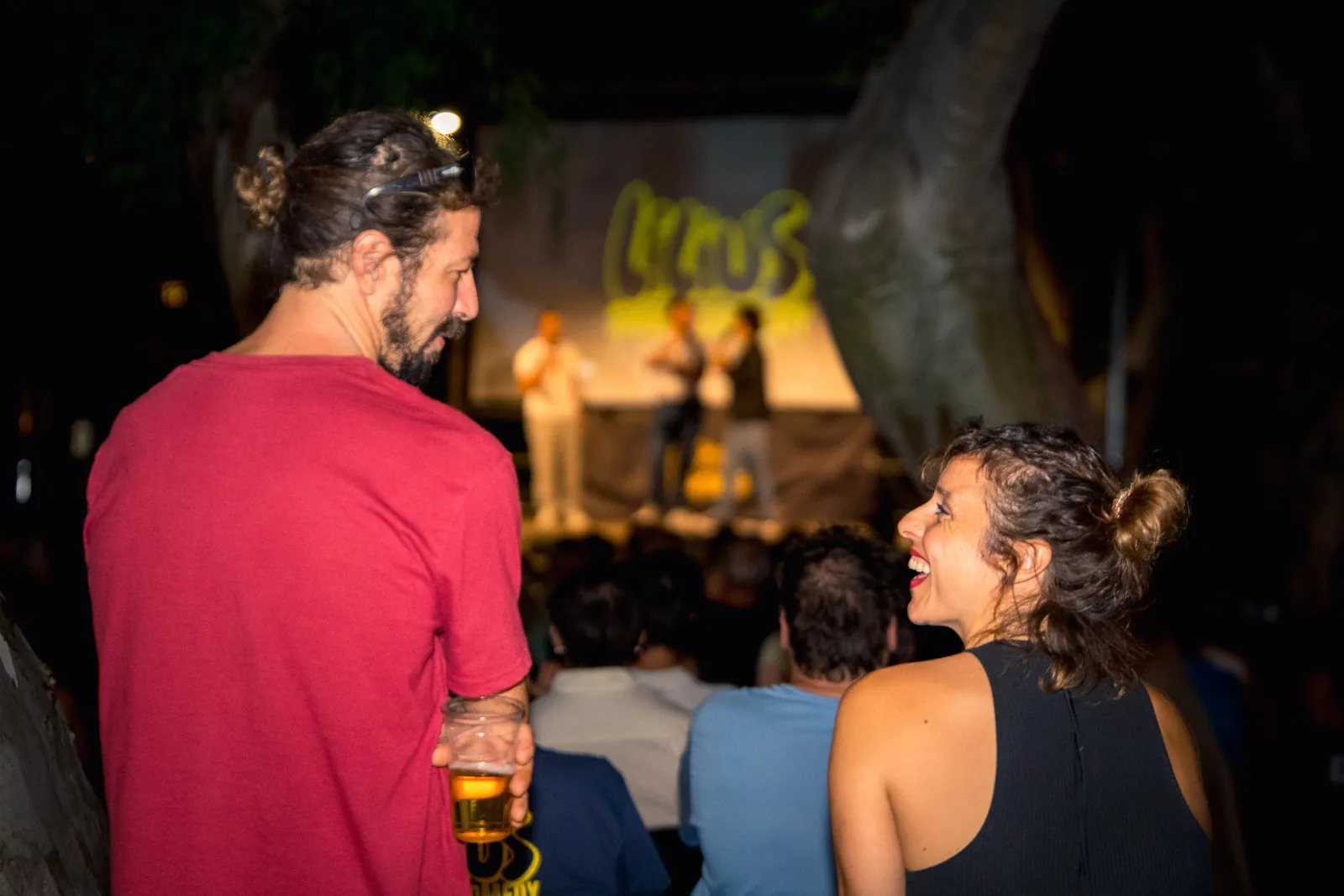 Una coppia di spettatori ripresa di spalle mentre chiacchiera, con il palco e lo schermo sfocati sullo sfondo tra gli alberi