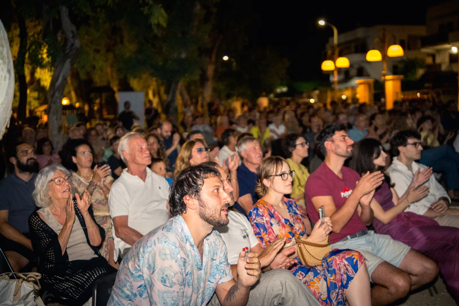 Vista ampia del pubblico seduto tra gli alberi, tra luci calde e applausi