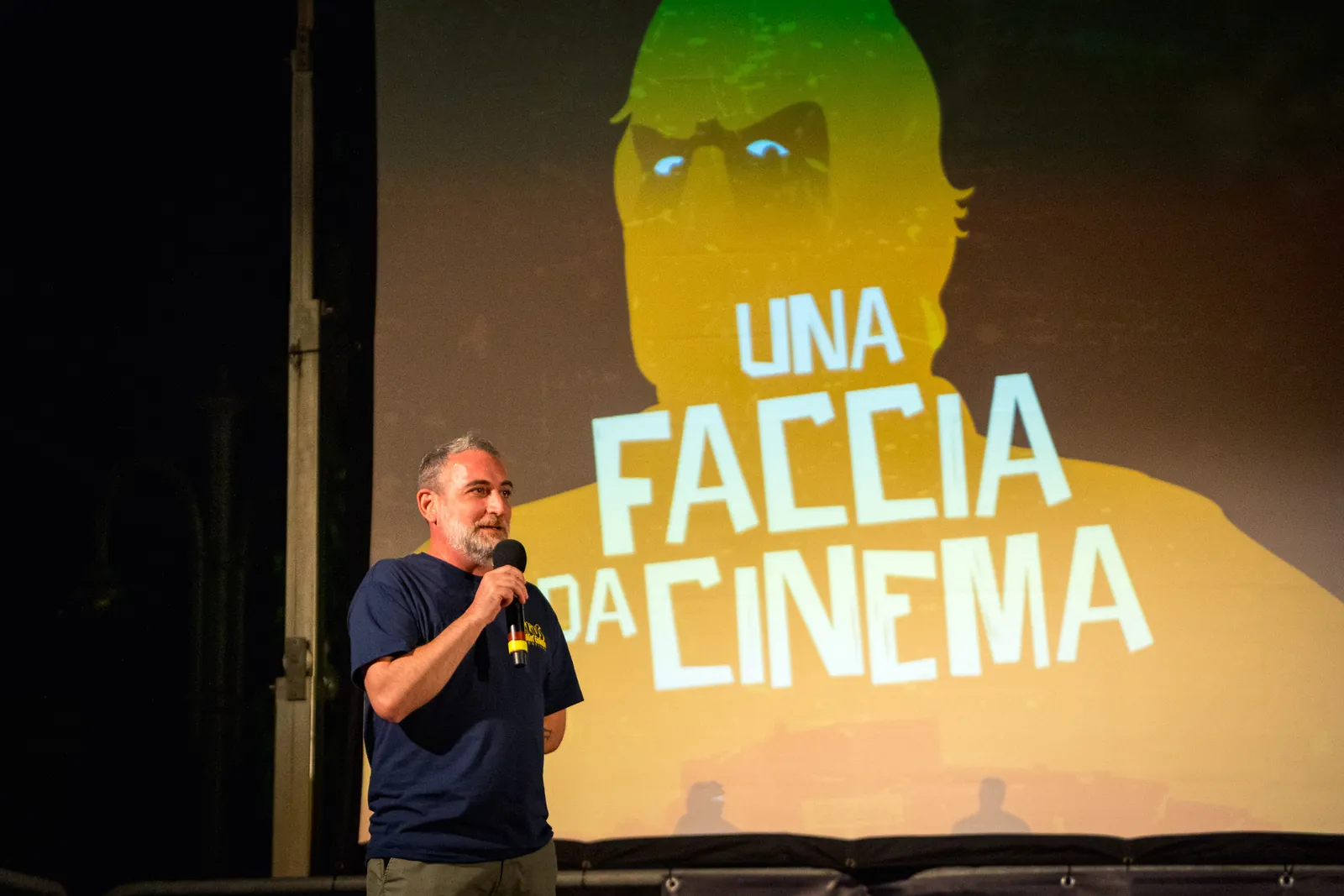 Un uomo con il microfono sul palco, davanti a un personaggio animato verde e minaccioso proiettato sullo schermo alle spalle