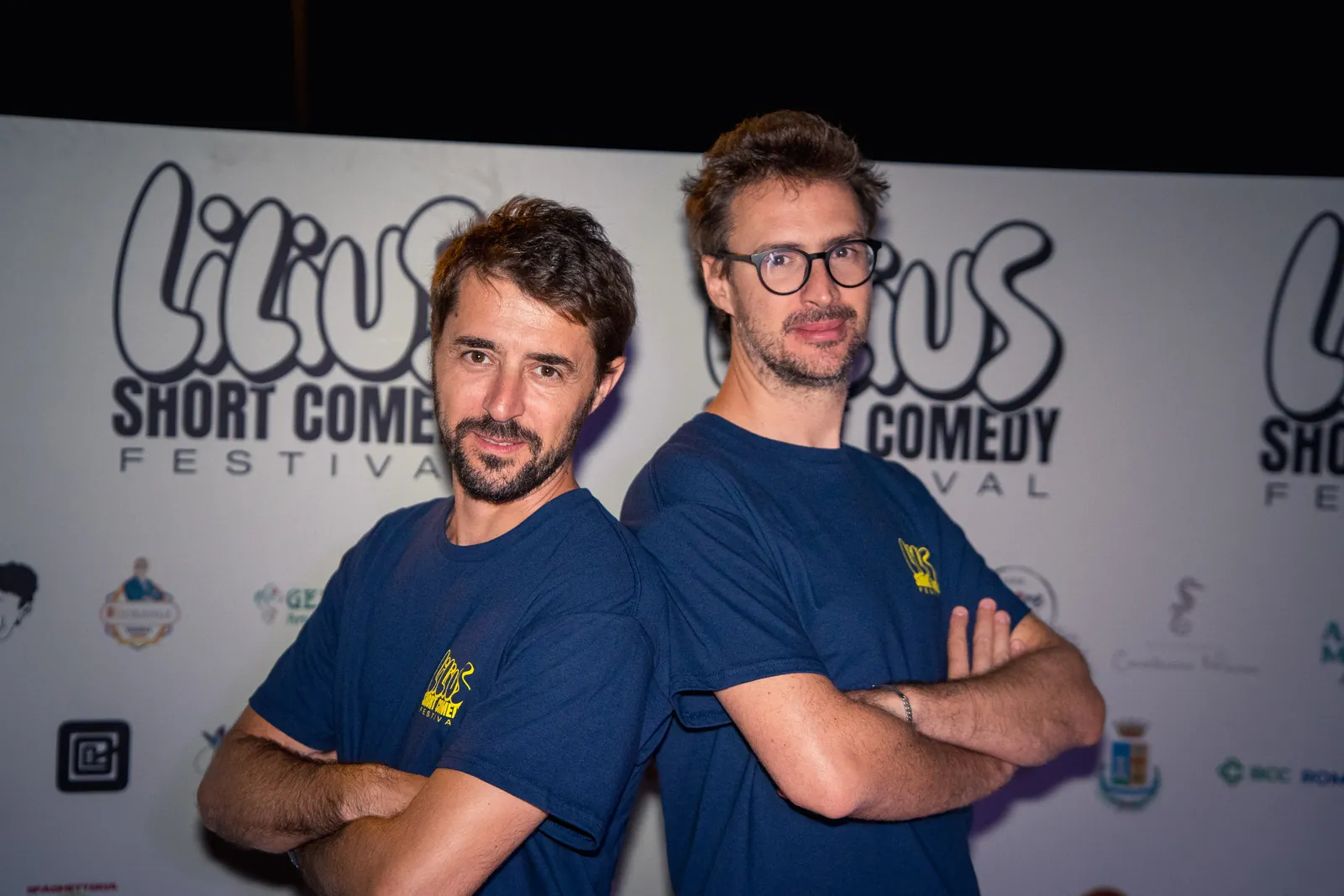 Due uomini in maglietta blu del festival posano a braccia conserte davanti al backdrop