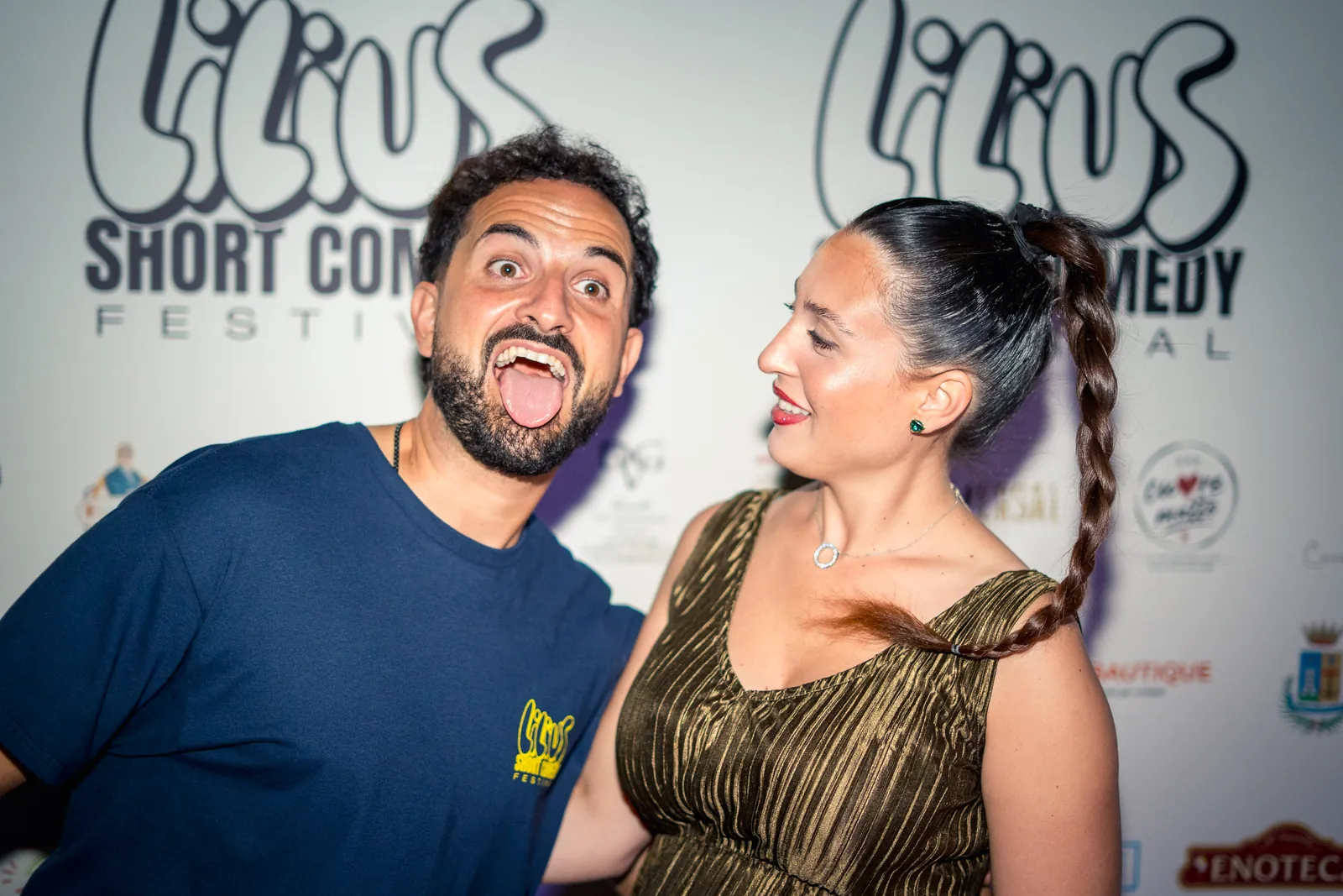Un uomo fa la linguaccia mentre una donna ride, faccia a faccia davanti al backdrop del festival