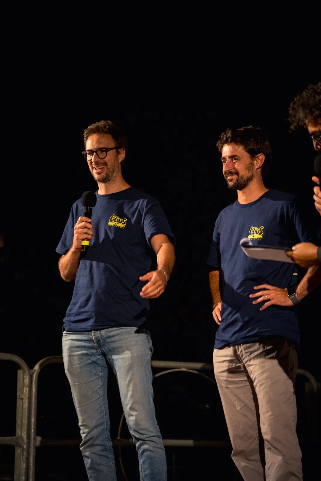 Due uomini in maglietta blu del festival sul palco: uno tiene il microfono e parla, l'altro ascolta