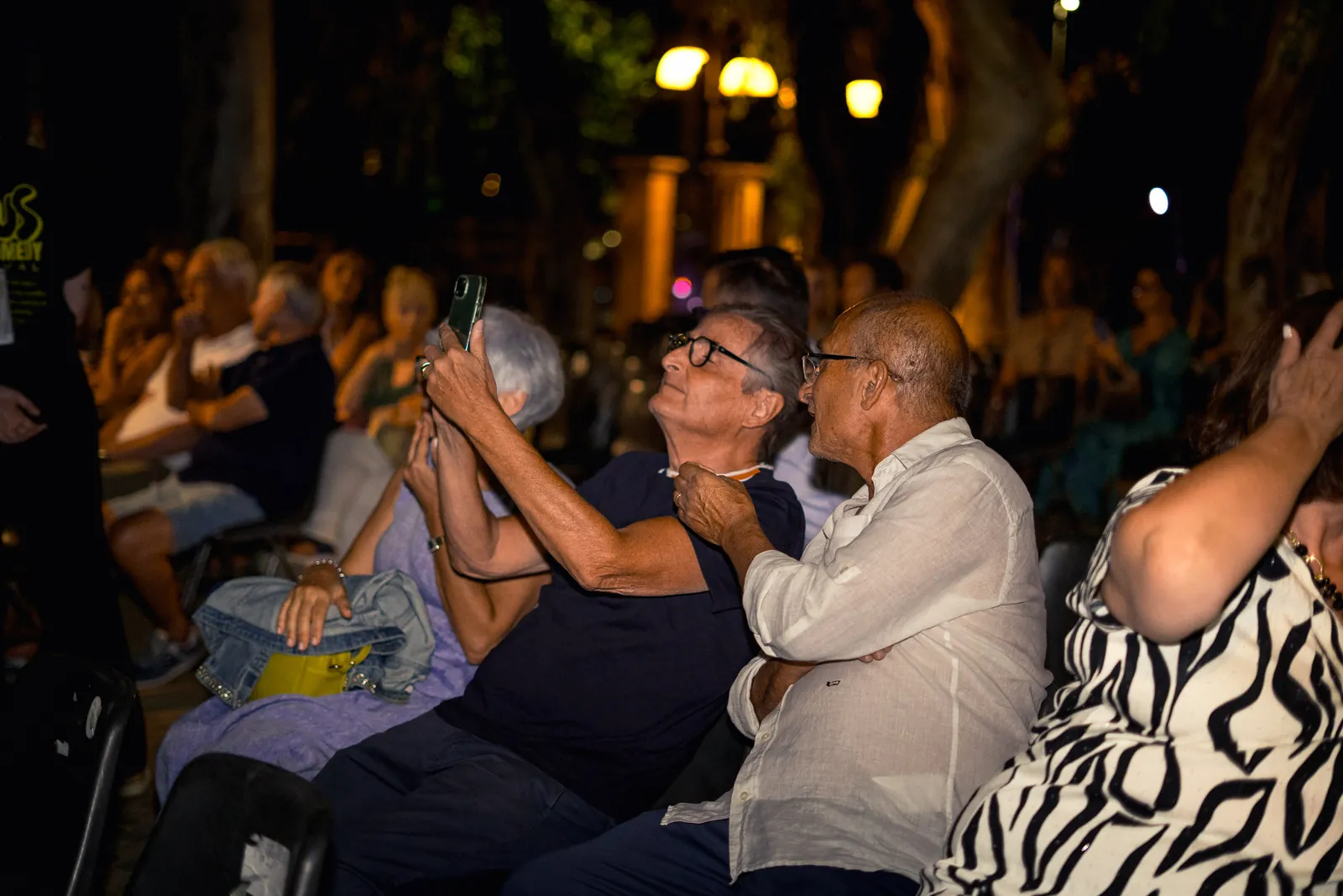 Il pubblico applaude, con una coppia di spettatori anziani in primo piano sotto le luci della sera