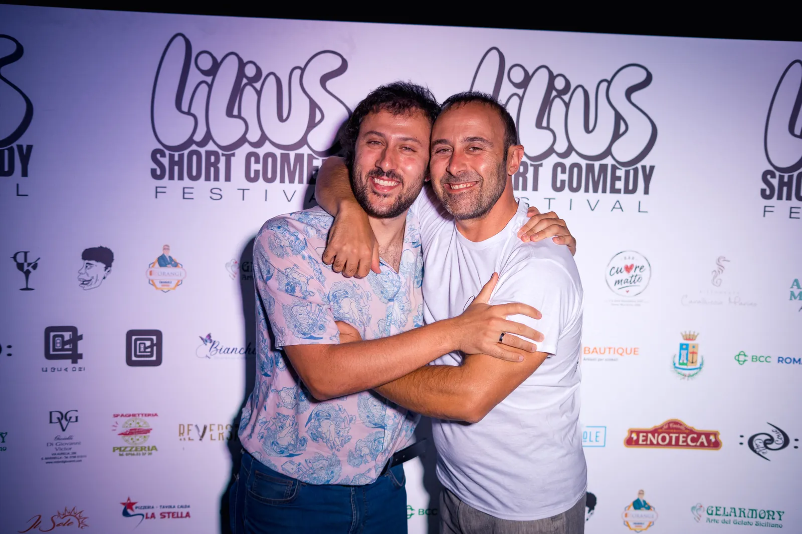 Due amici abbracciati e sorridenti davanti al backdrop del festival