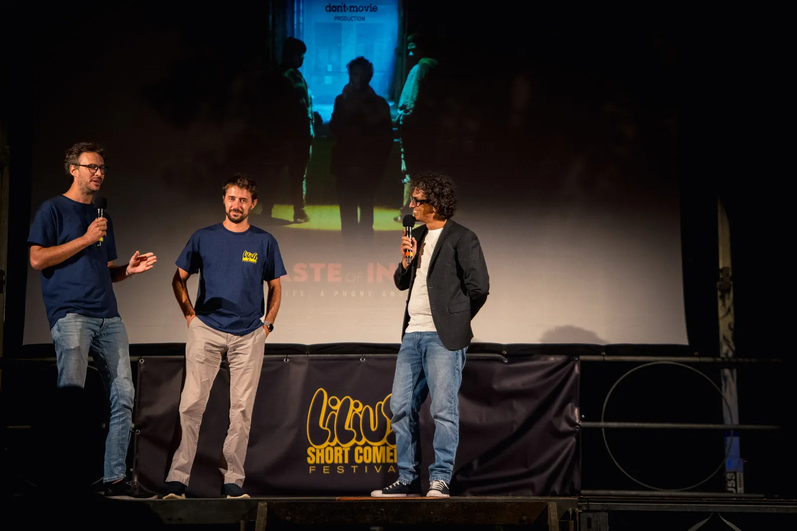 Tre persone sul palco con i microfoni, con una sagoma proiettata sullo schermo alle spalle