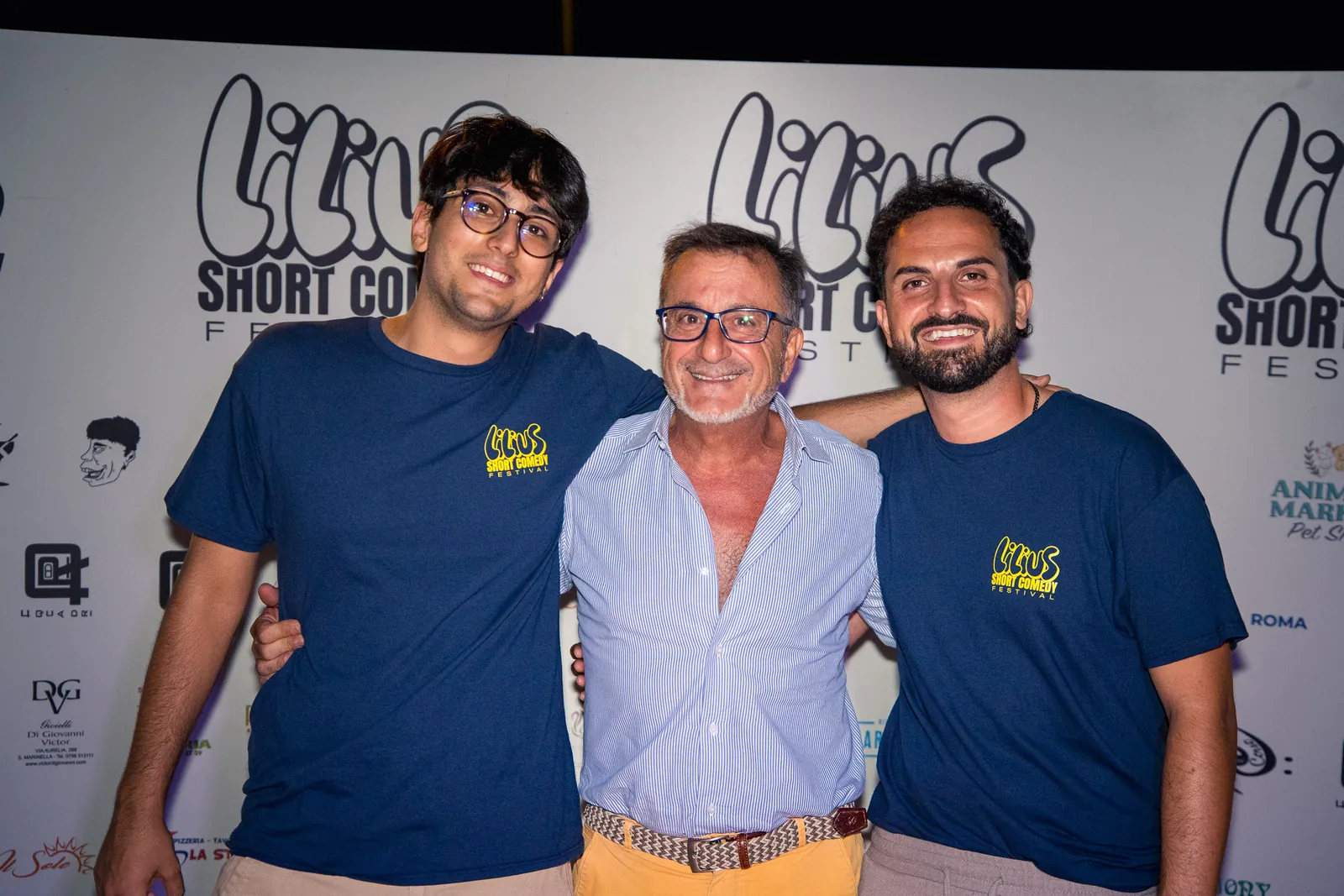 Tre amici abbracciati e sorridenti al photo-wall, uno con camicia chiara e cintura vistosa