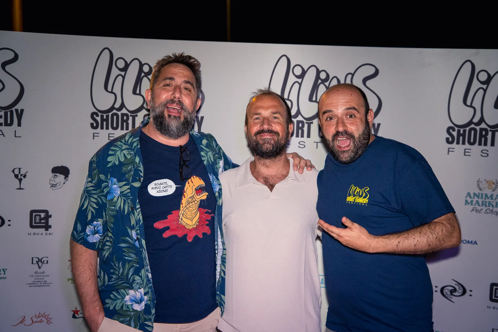 Tre amici in posa scherzosa al photo-wall, uno con camicia a fiori tropicale