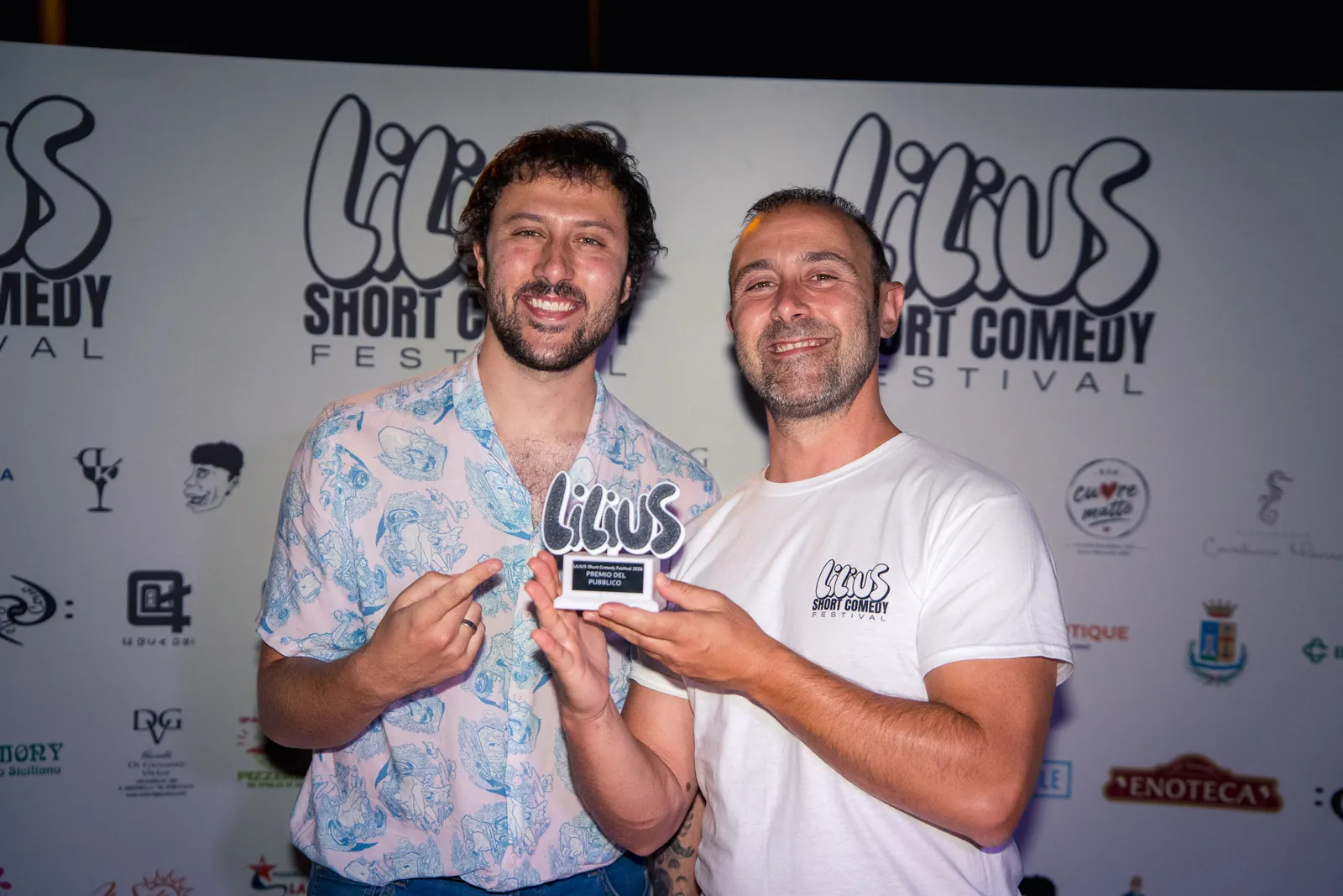 Due vincitori sorridenti mostrano insieme il trofeo del Premio del Pubblico davanti al photo-wall del festival