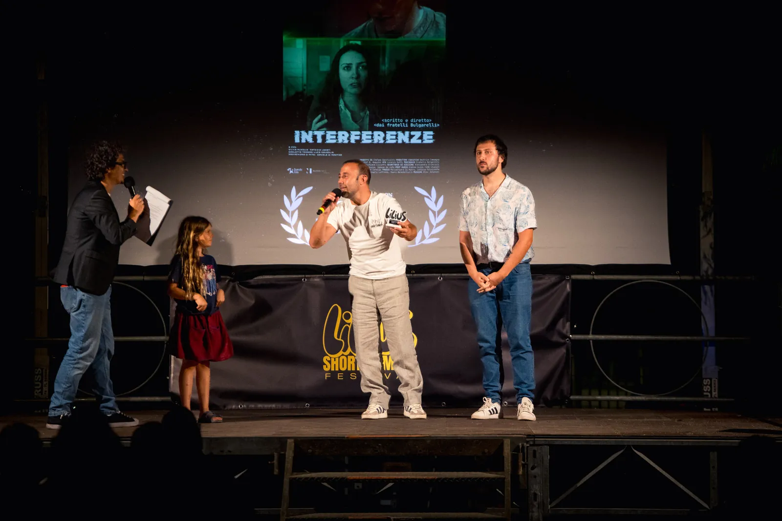 Sul palco, un uomo con microfono e appunti in mano, una bambina al suo fianco, e altri due uomini davanti alla locandina del cortometraggio Interferenze incorniciata da corone d'alloro, proiettata sullo schermo