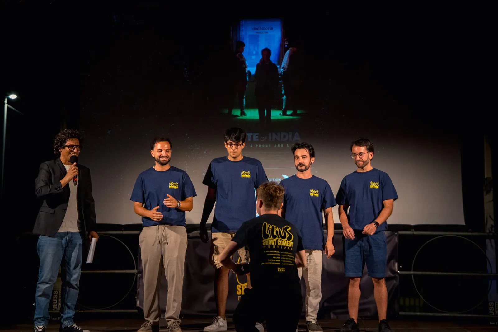 Sul palco, i quattro membri della squadra di Taste of India in maglietta blu del festival, con un uomo al microfono e la locandina del film proiettata alle spalle